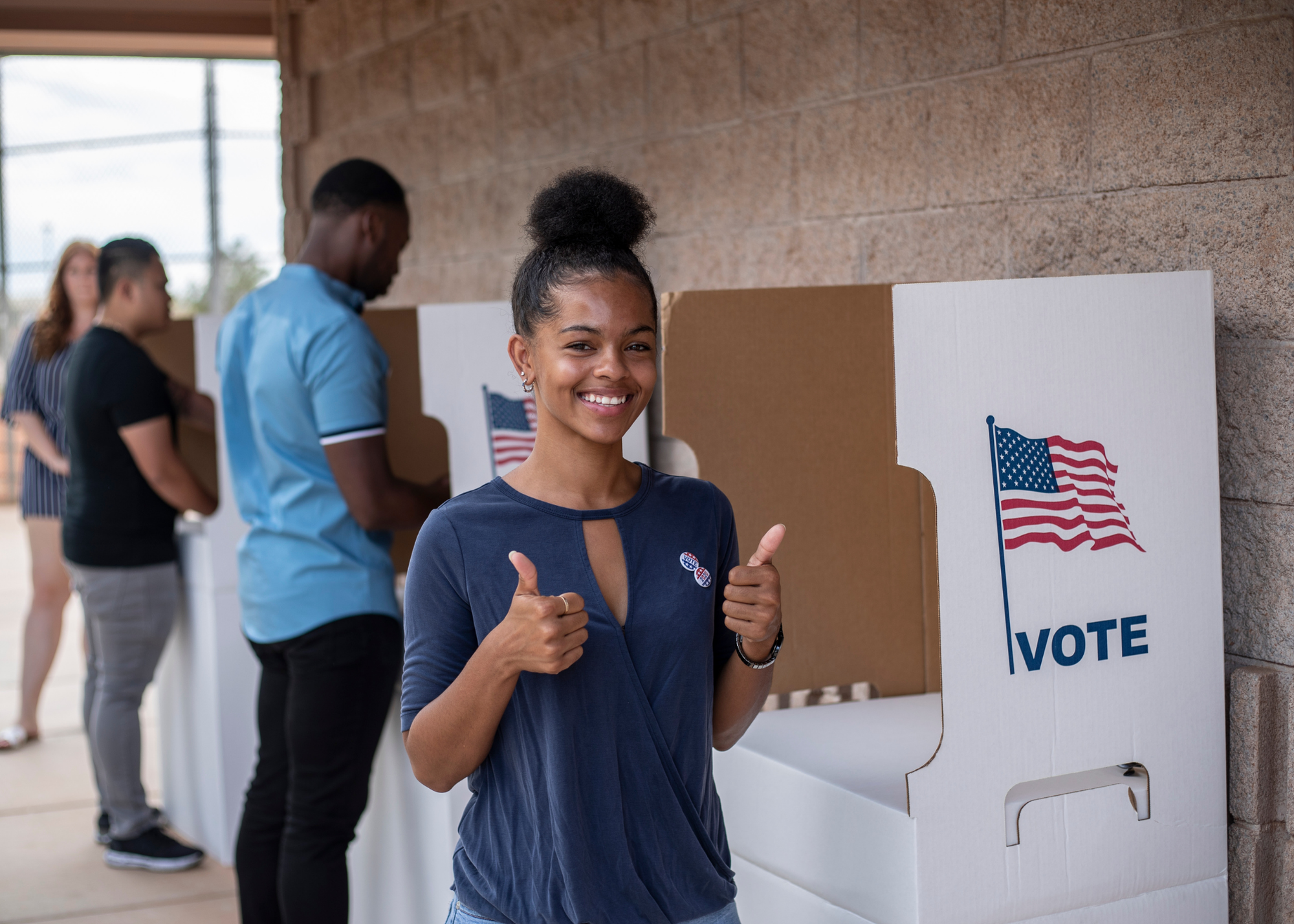 Smiling young woman with natural hair in a bun giving a thumbs up at a voting booth, with other voters in line, American flags on voting signs.