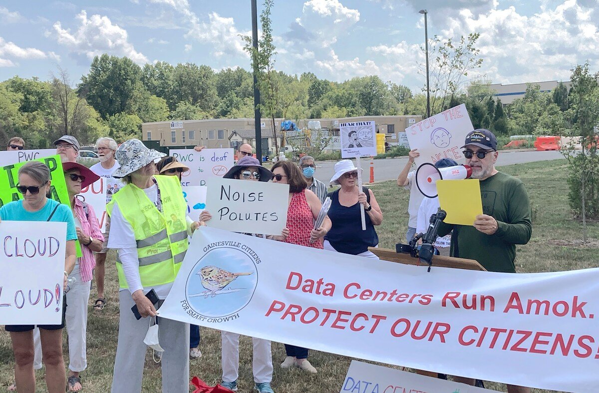 Group of people protesting outdoors with signs about noise pollution and data centers, some wearing hats and sunglasses.
