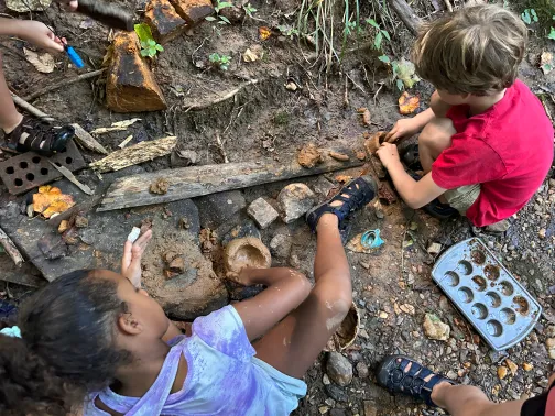 Two children playing with mud and rocks outdoors, one girl lying down and one boy sitting, surrounded by natural elements.