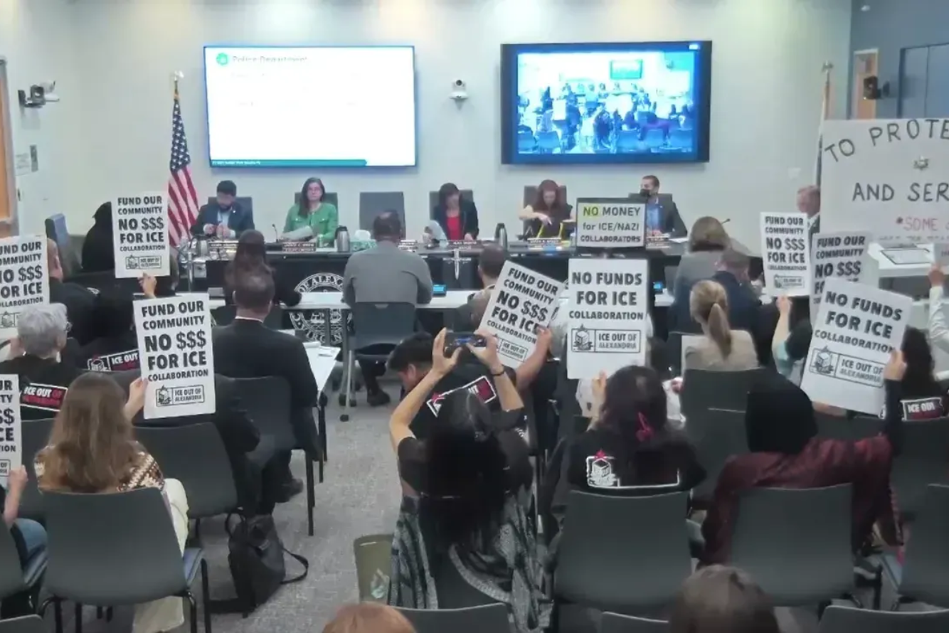 A city hall meeting with protesters holding signs that say 'FUND OUR COMMUNITY NO $$ FOR ICE' and 'NO FUNDS FOR ICE.' Several people are seated at a long panel table at the front of the room, with two large screens behind them. The American flag stands next to the panel.