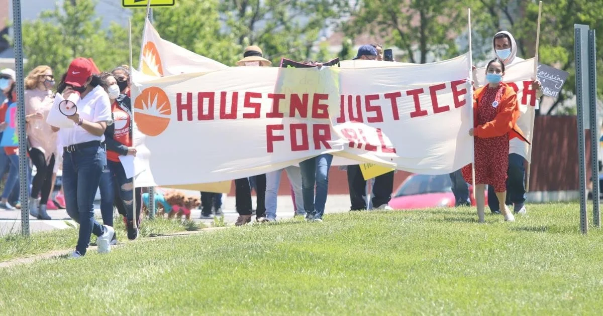 Group of protesters holding a large banner that reads 'Housing Justice For All' during a demonstration on a sunny day.