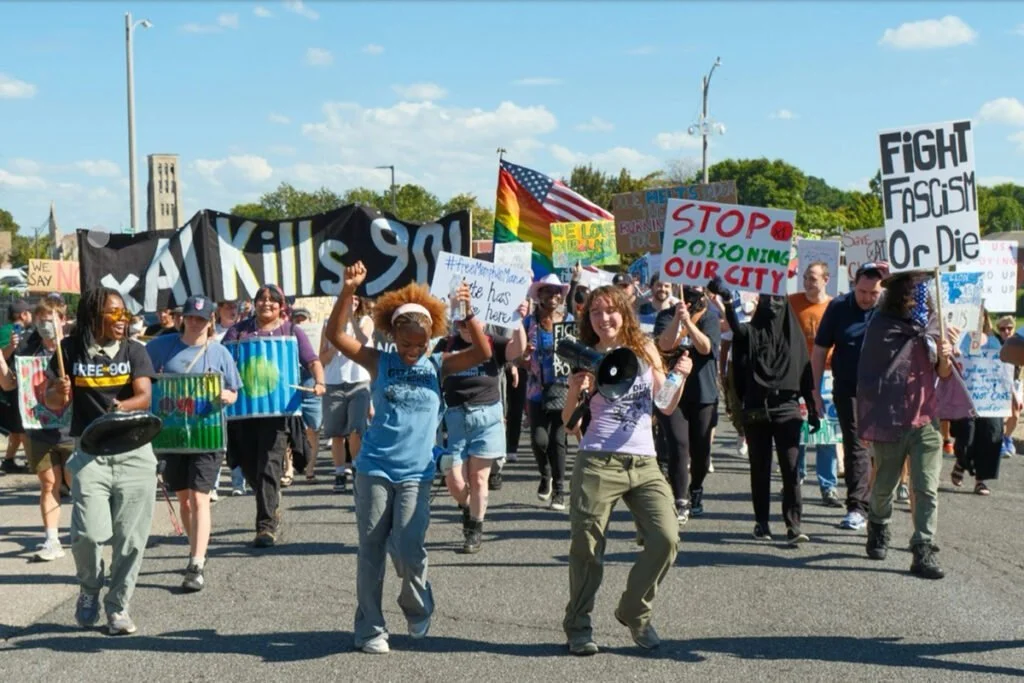 People participating in a protest march holding signs and banners advocating against racism and fascism, with some signs reading 'Kill 90', 'Stop Poisoning Our City', and 'Fight Fascism or Die', under a partly cloudy sky.