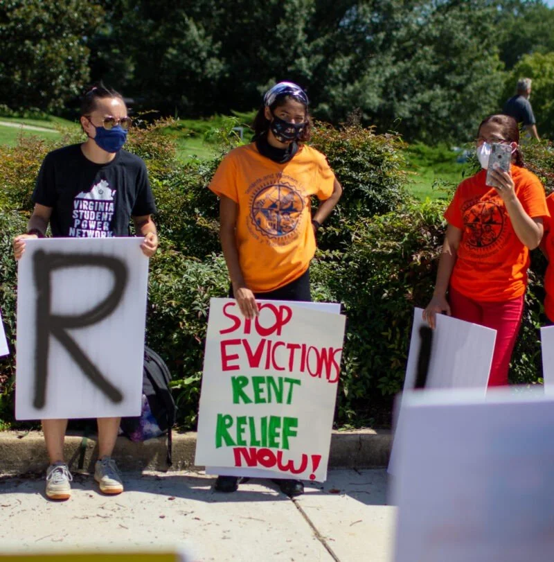 Protesters wearing face masks holding signs advocating rent relief and stopping evictions in a park with trees in the background.
