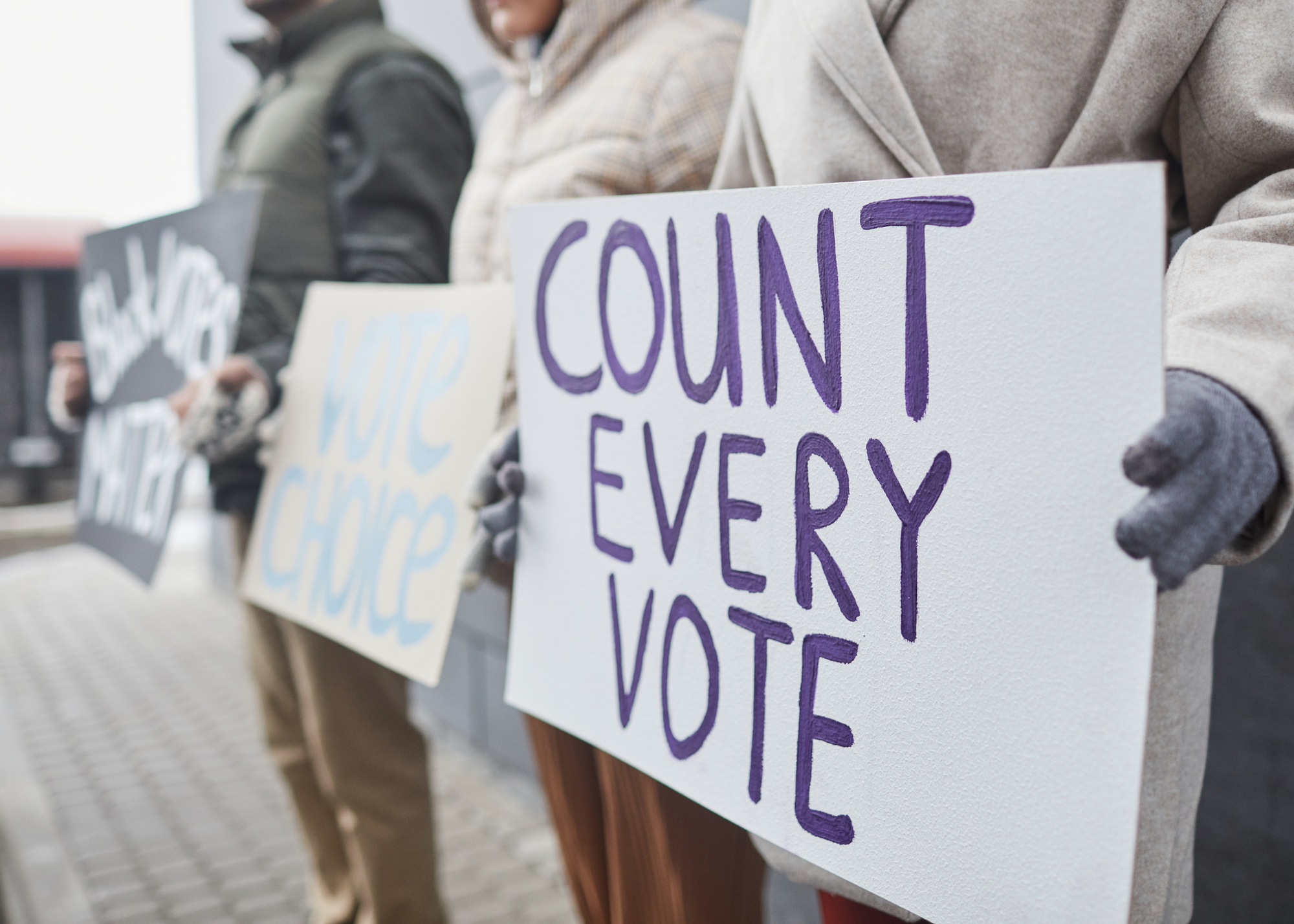 People holding protest signs, one with purple text reading 'Count Every Vote'.