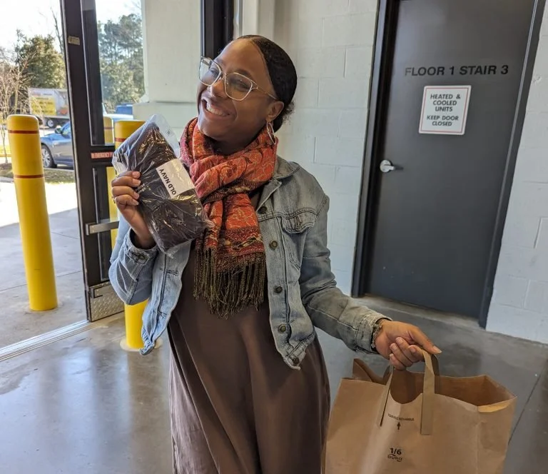 A woman smiling inside a store, holding a packaged item in one hand and a paper shopping bag in the other, near an exit door with a sign.