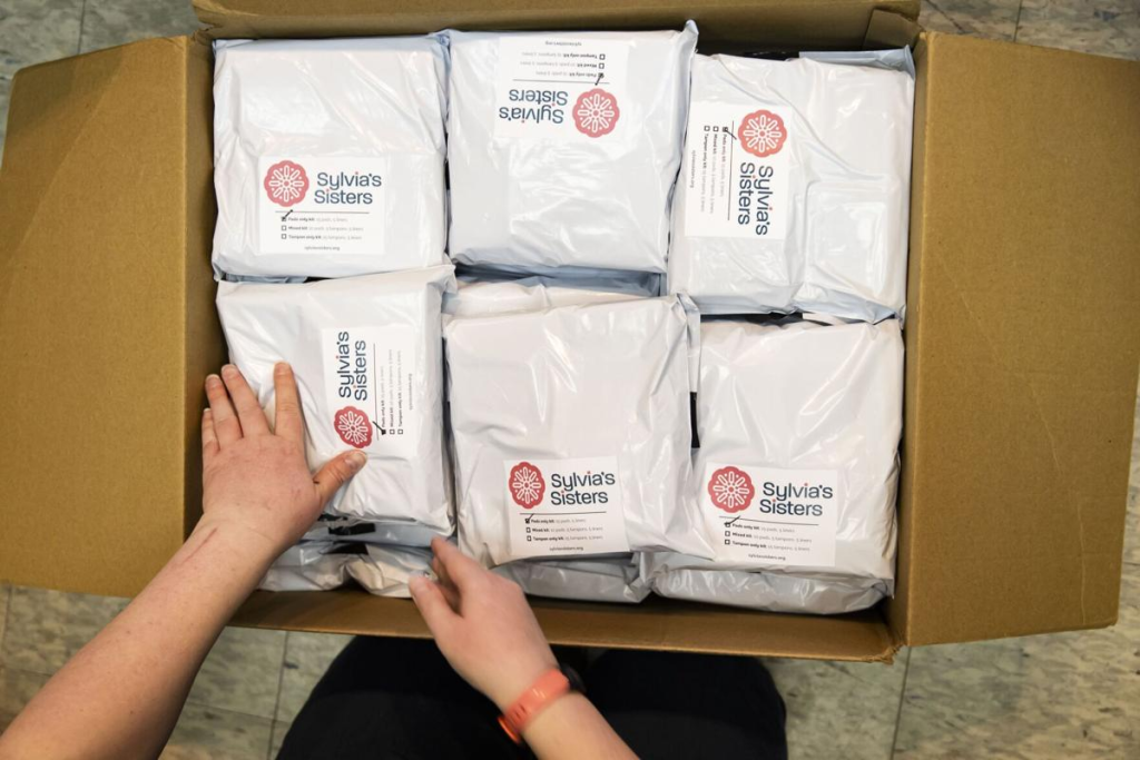 A person opening a cardboard box containing six packages wrapped in white plastic with labels for Sylvia's Sisters.