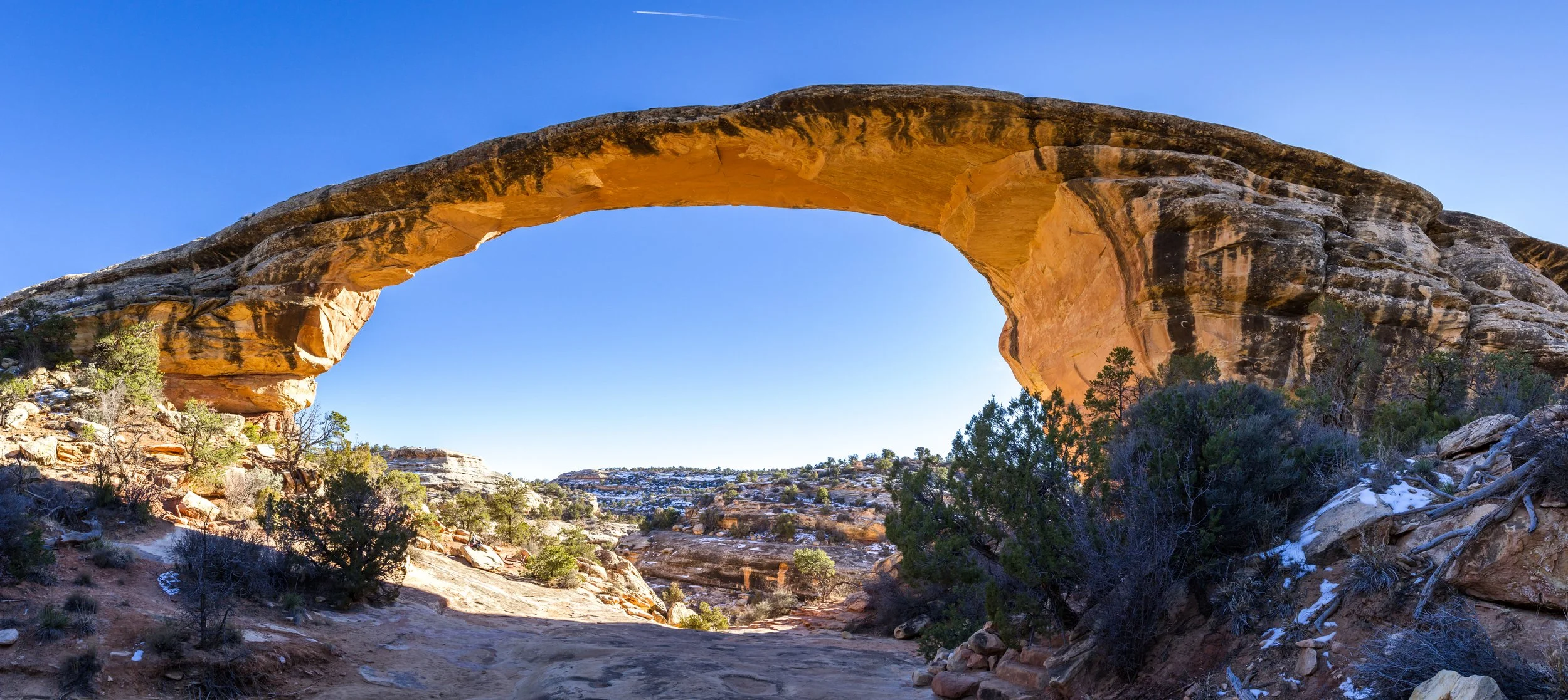 A natural stone arch in a desert landscape with sparse vegetation and distant hills, under a clear blue sky.