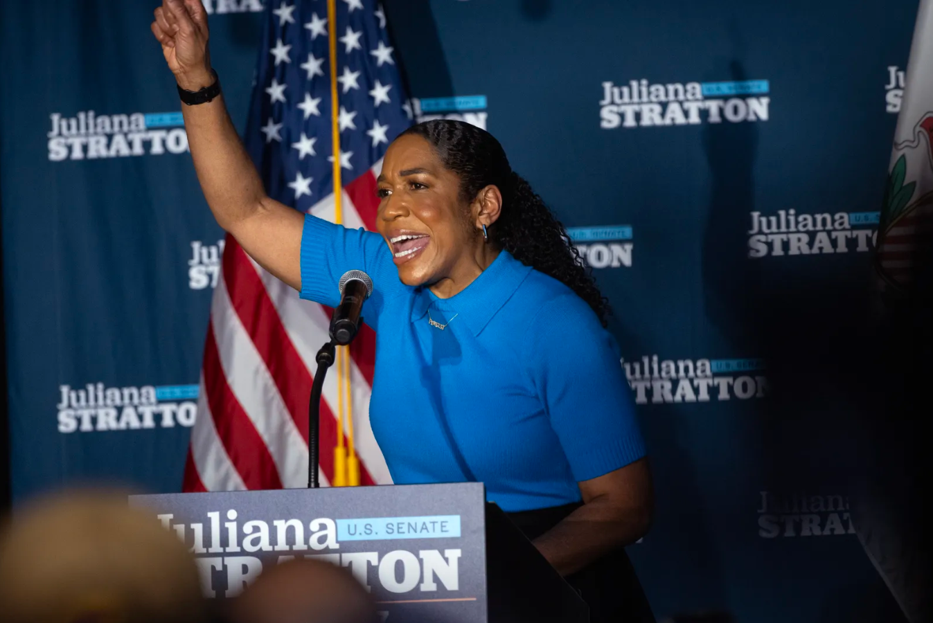 A woman in a blue shirt speaking at a campaign event, raising her right arm. Behind her is a U.S. flag and a blue background with the text 'Juliana Stratton'.