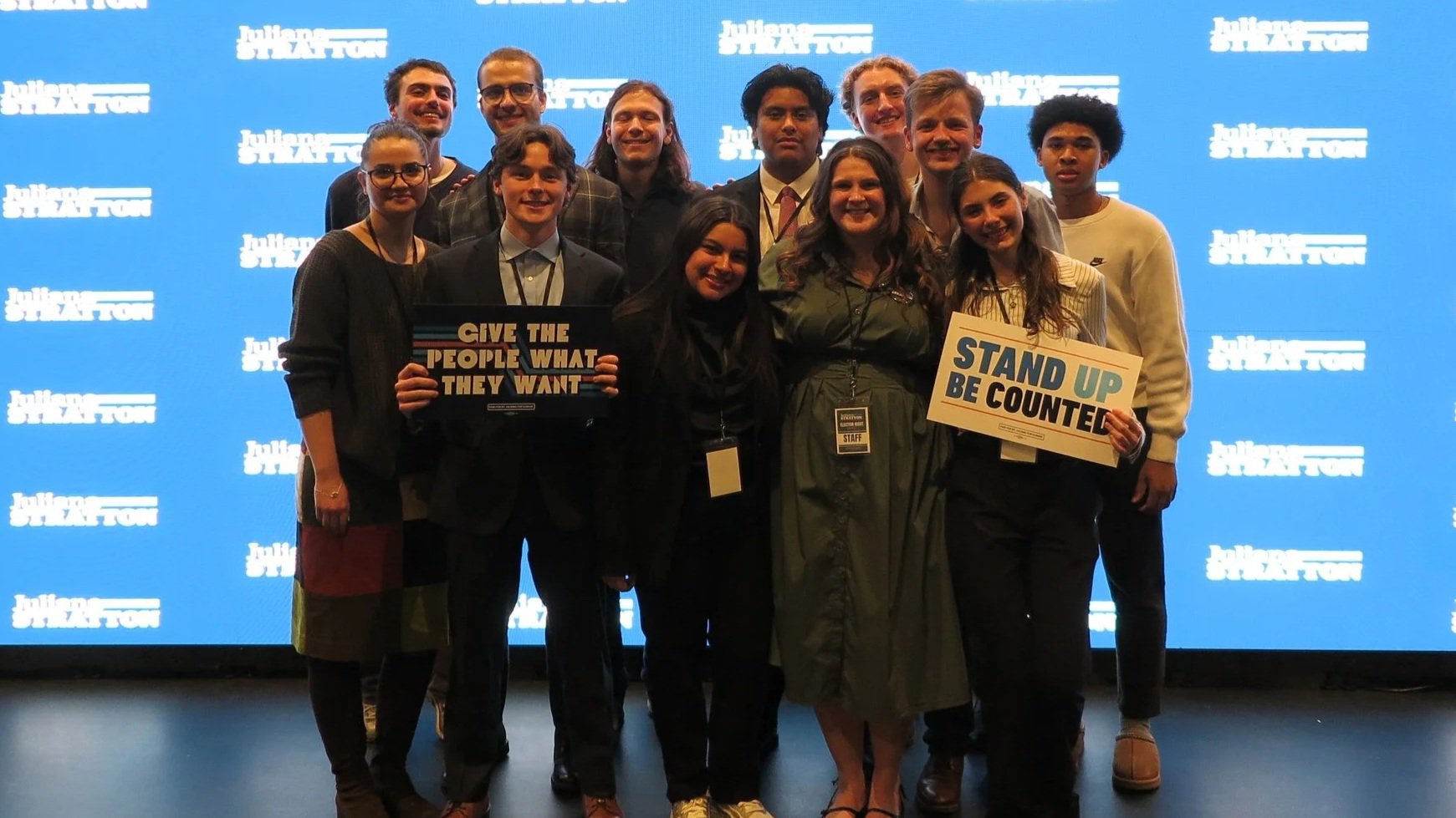 Group of young adults and staff at an event with a blue background displaying white text that says 'Julius G. W. F. L'. Two people are holding signs reading 'GIVE THE PEOPLE WHAT THEY WANT' and 'STAND UP BE COUNTED'.