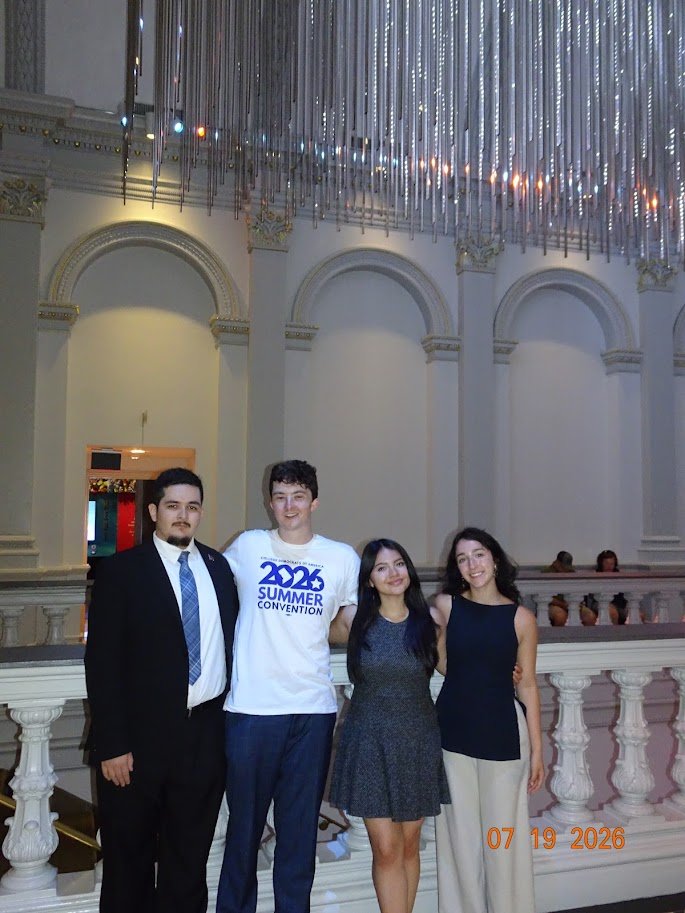Four young adults, three women and one man, are standing together inside a grand, elegant hall with high arched ceilings and decorative moldings. They are smiling and posing for a photo. The ceiling is decorated with hanging metallic streamers or ribbons, and the background features a white balustrade with decorative urns. The photo was taken on July 19, 2026.