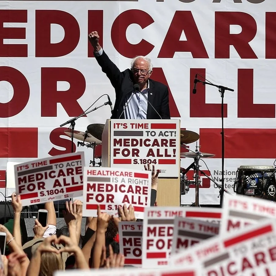 Bernie Sanders speaking at a rally with a large crowd holding signs that read 'Medicare for All' and 'It's Time to Act!'.
