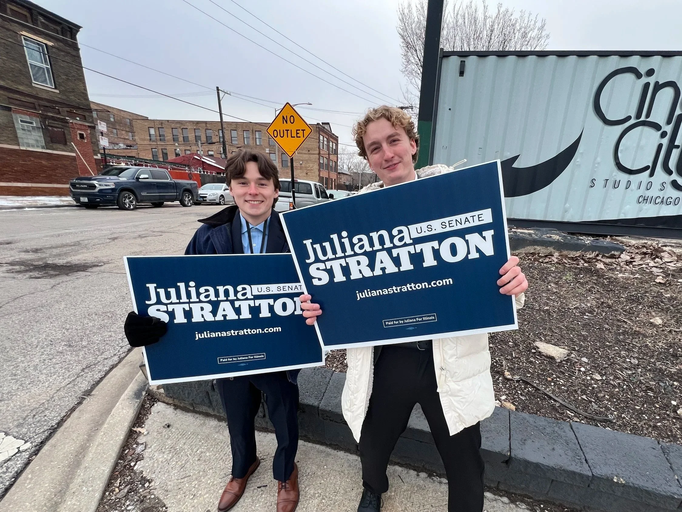 Two young people protesting, holding signs that read 'Juliana Stratton U.S. Senate.' They are standing outdoors on a city street, with buildings, cars, and a large sign for 'Studio Chicago' visible in the background.