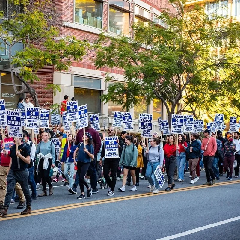 A large group of people participating in a protest march on a city street, holding signs that say 'UAW ON STRIKE' and 'UNFAIR LABOR PRACTICE,' with buildings and trees in the background.