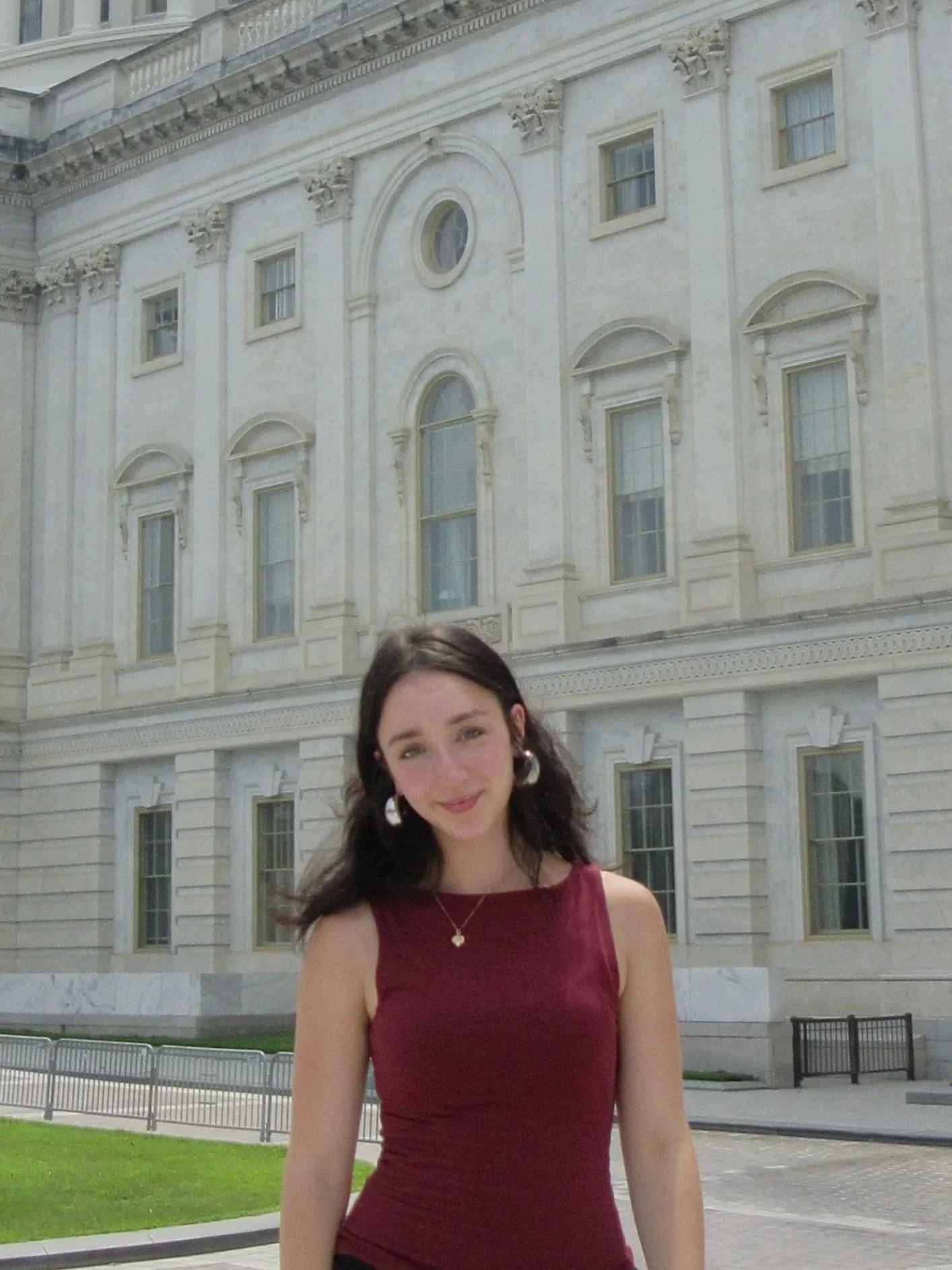 A young woman with long dark hair and earrings, wearing a maroon sleeveless dress, standing in front of a white ornate building with multiple windows and decorative architectural details.