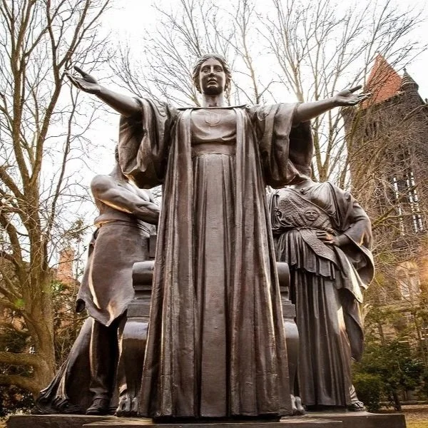 Statue of a woman with arms outstretched, surrounded by smaller figures, in an outdoor park setting with trees and buildings in the background.