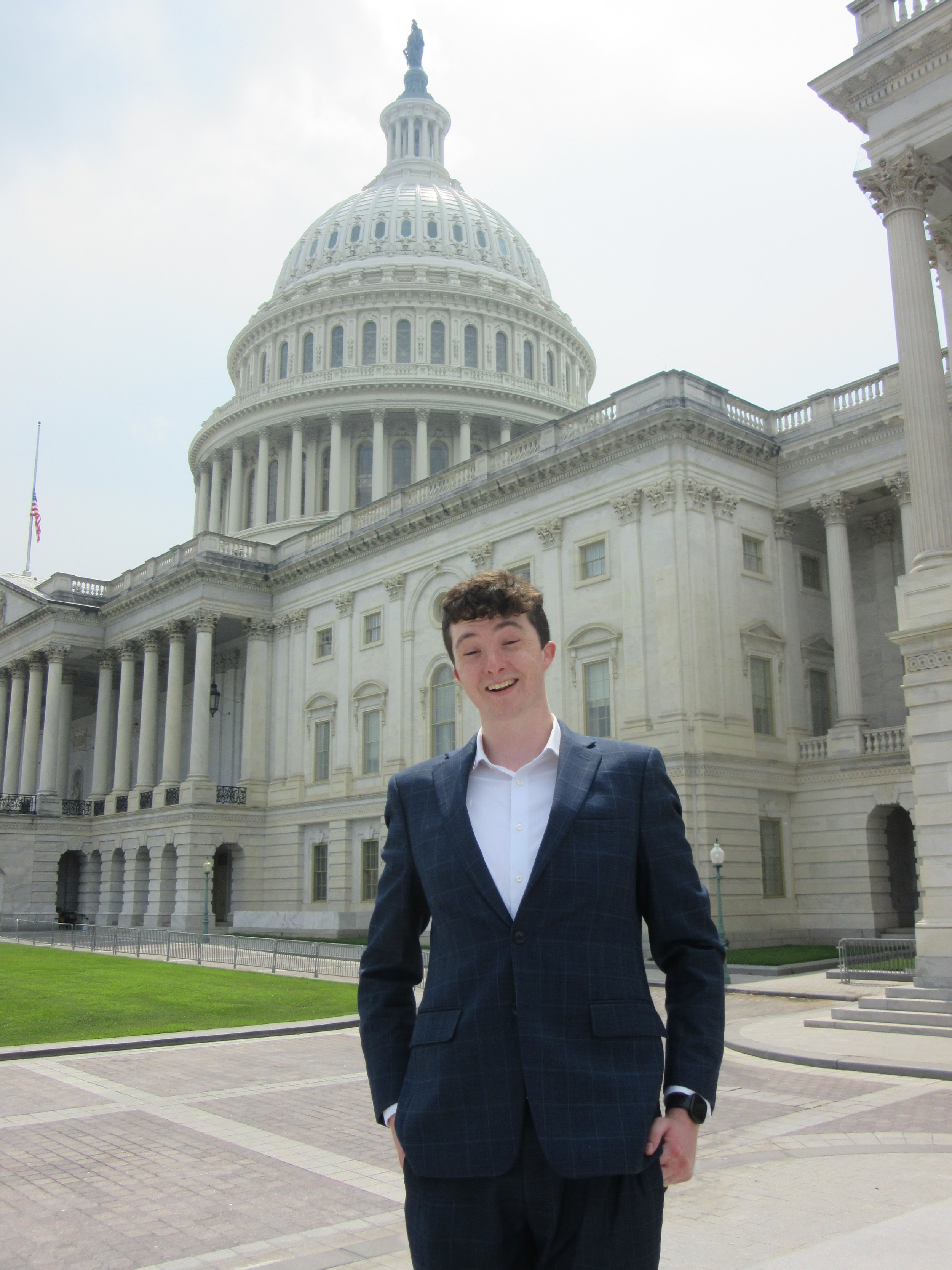 A young man in a suit standing outside the U.S. Capitol building with a large dome, columns, and stairs, smiling at the camera.