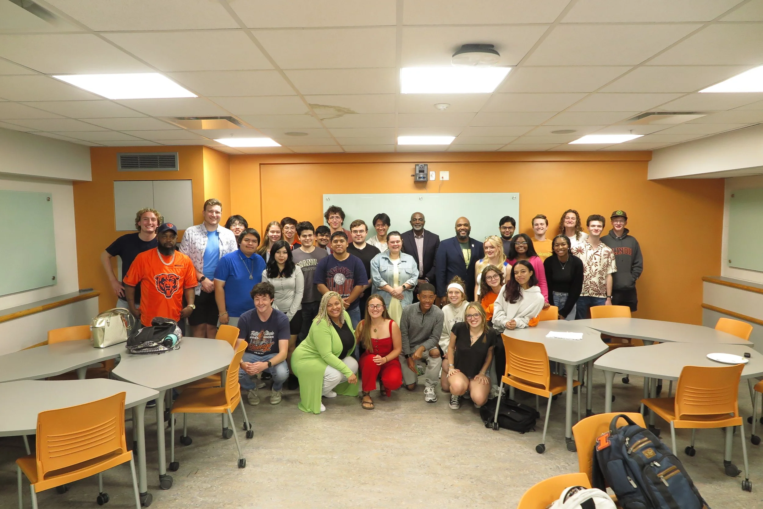 A large group of people, including young adults and a few older individuals, gathered in a classroom with orange and white walls. The group is standing and sitting in front of a whiteboard, smiling at the camera. The classroom has round tables with orange chairs, some with backpacks and personal items on them. The ceiling has fluorescent lights, and there's a whiteboard on the wall behind the group.