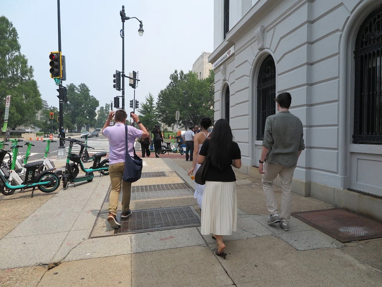 People walking on a city sidewalk near a white building, with a green bike-sharing station and traffic lights at an intersection, during daytime.