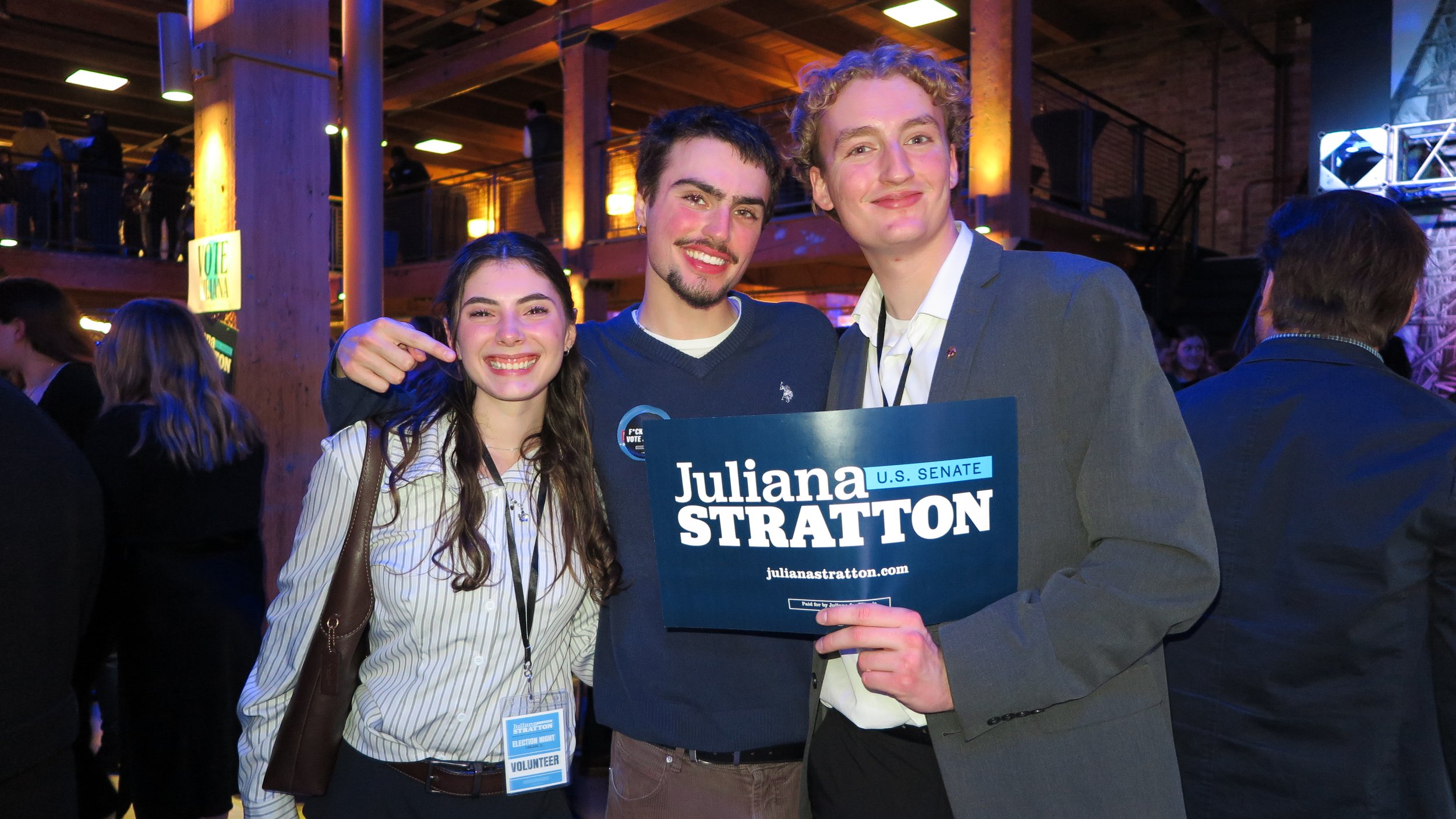 Three young adults smiling and posing for a photo at an indoor event. The man on the right is holding a sign that reads 'Juliana STRATTON U.S. SENATE' and has a campaign website. The woman on the left is wearing a striped shirt, and the man in the middle is dressed in a dark sweater. They are surrounded by other people in a warmly lit, rustic setting with wood beams and hanging lights.