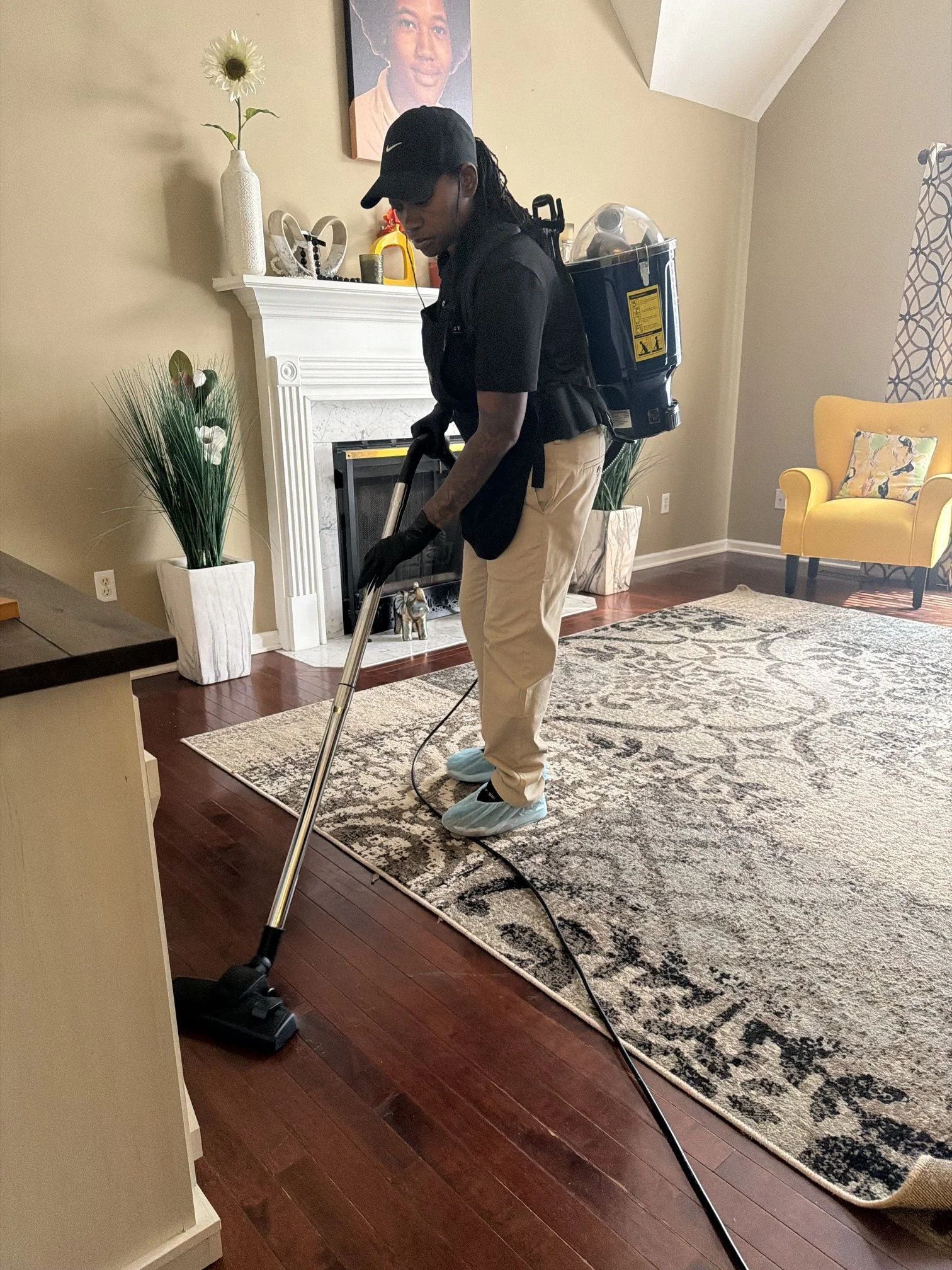 A person wearing a black polo shirt, tan pants, black gloves, disposable shoe covers, and a black cap, is vacuum cleaning a living room area with a patterned rug, a yellow armchair with a bird-patterned pillow, and green plants. They are equipped wit