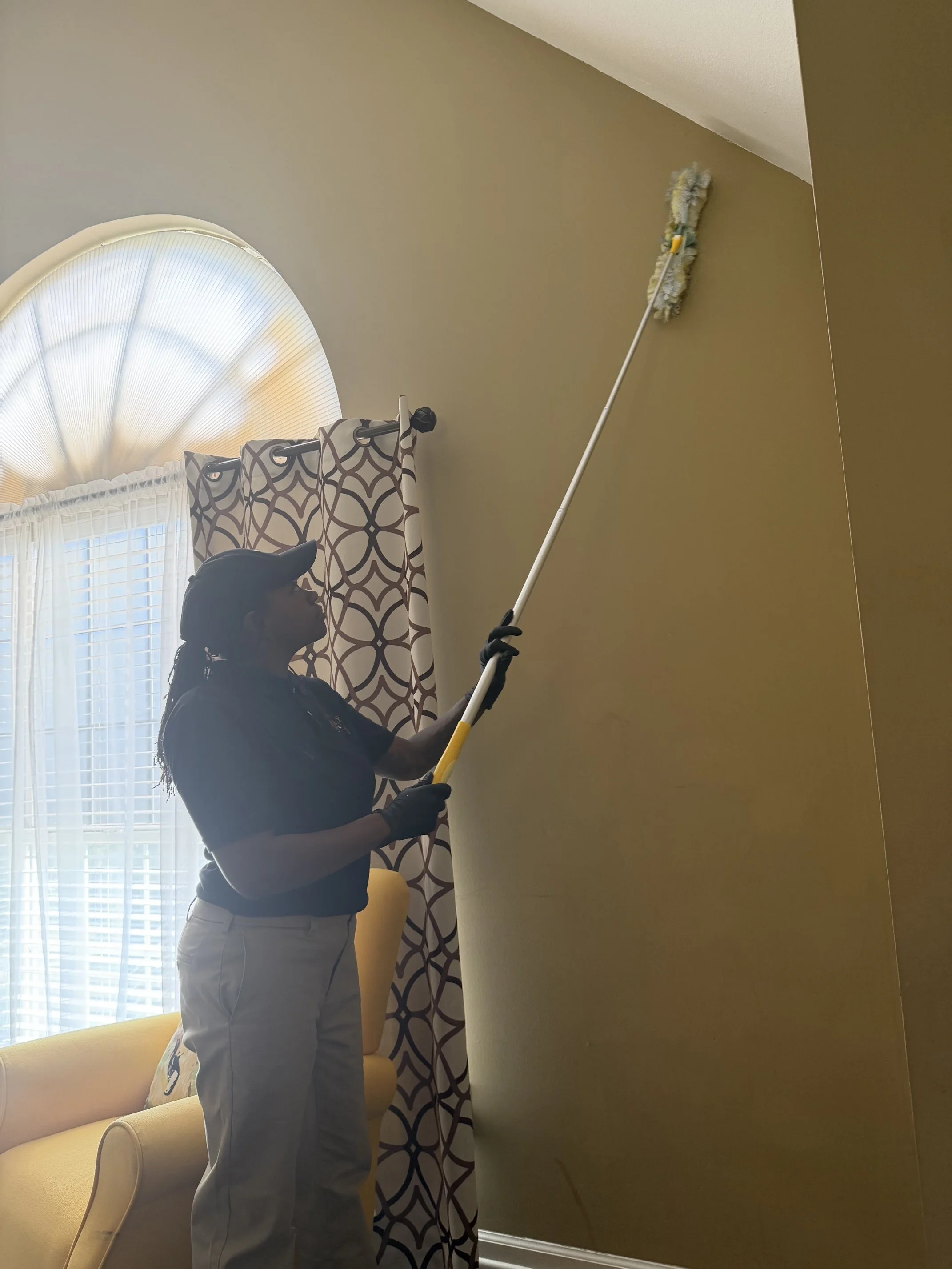 A person in a uniform and cap is cleaning a high wall with a long-handled duster in a room with patterned curtains and a yellow chair.