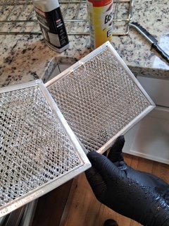 Person wearing a black glove holding a metallic mesh filter, with cleaning supplies in the background on a kitchen countertop.