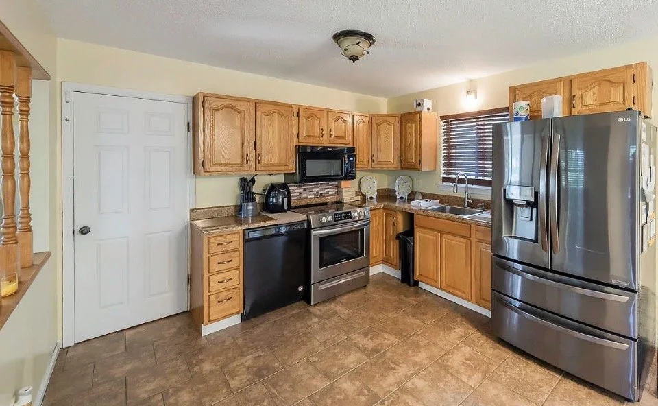 Kitchen with wooden cabinets, stainless steel refrigerator, oven, microwave, and a window above the sink.