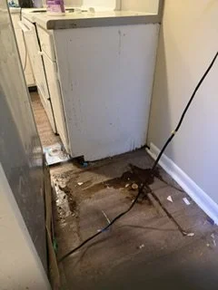 Damaged hardwood floor near a kitchen wall with exposed subfloor and a loose electrical cord.