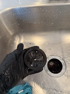 A person's hand in a black glove holding a black drain strainer over a kitchen sink with water droplets on the sink wall.