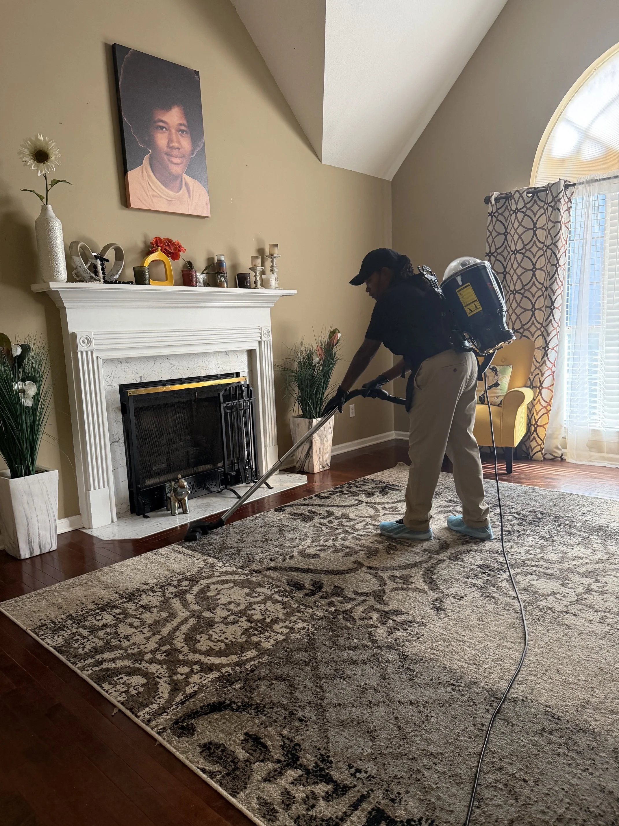 A person in protective gear uses a fogging device in a living room with a fireplace, large rug, and decorative items on the mantel and plants on either side.