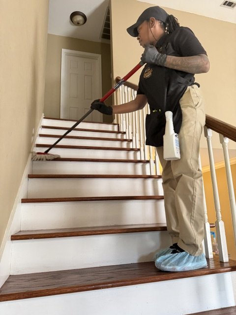 A woman wearing a black cap, black gloves, and a black t-shirt, mopping wooden stairs inside a house. She has a spray bottle hanging from her waist and is wearing blue shoe covers.