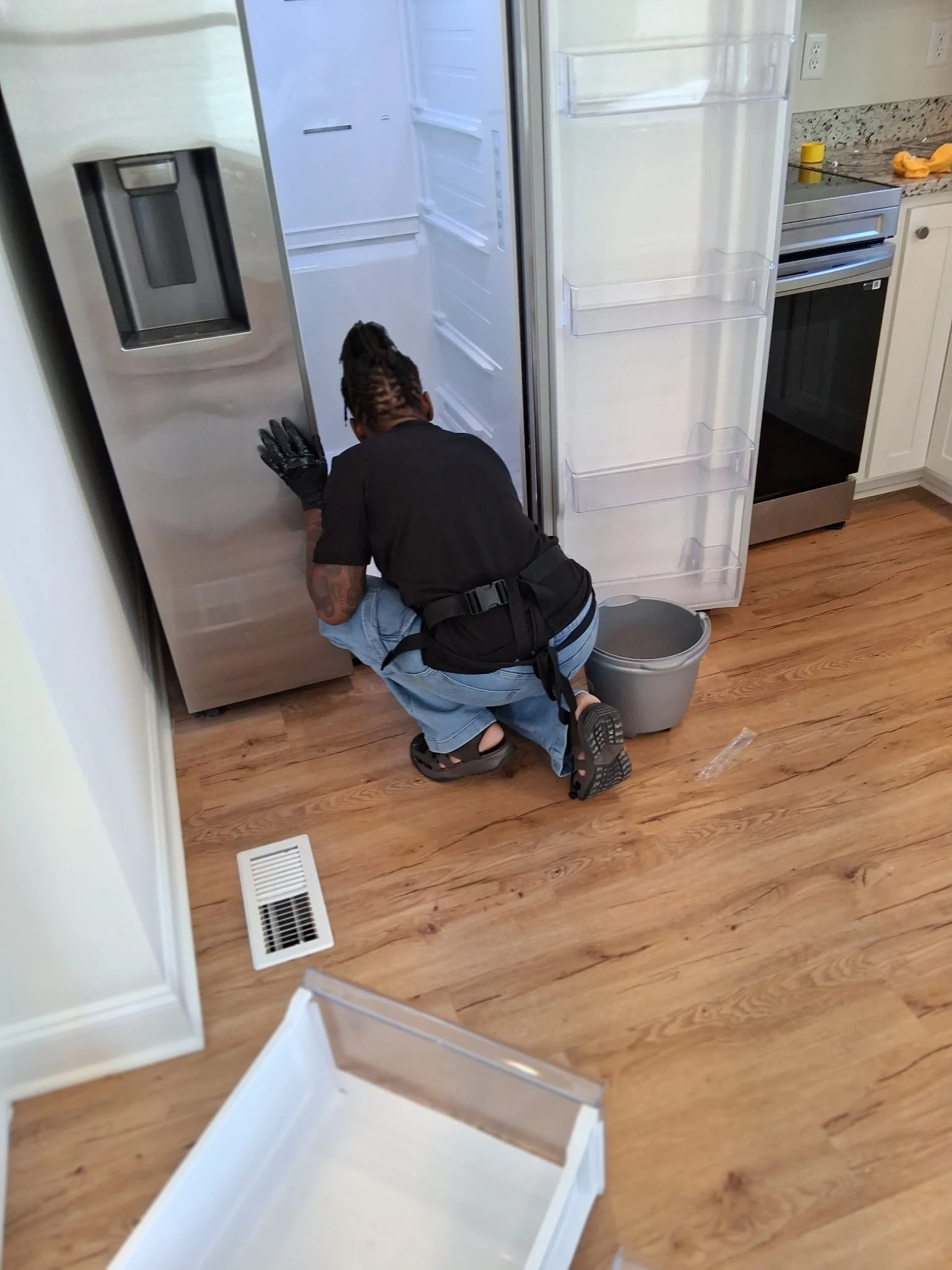 Person kneeling on wood floor cleaning inside an open empty refrigerator in a kitchen with white cabinetry and granite countertops.