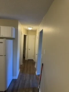Bright hallway with hardwood flooring, white walls, a closed door at the end, and a red broom leaning against the wall on the right.