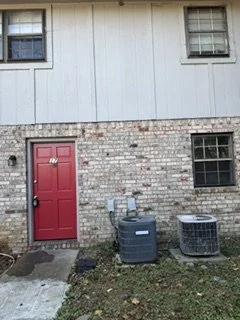 Exterior of a building with a red door, two small windows above, and two larger windows with black frames below, with grass and outdoor appliances near the door.