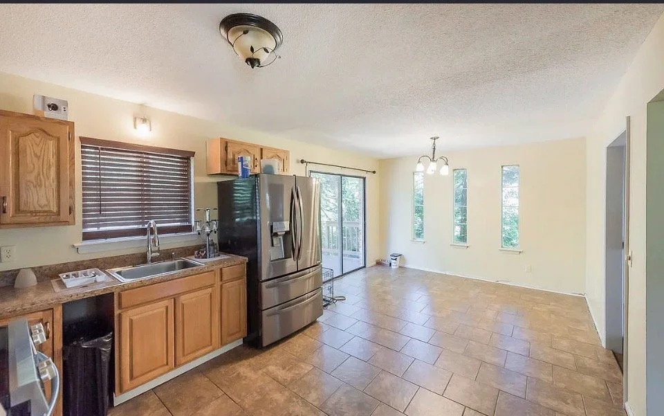 Kitchen with wooden cabinets, stainless steel refrigerator, window blinds, and a sliding glass door leading to an outdoor area. Adjacent dining area with three narrow windows, a chandelier, and tile flooring.