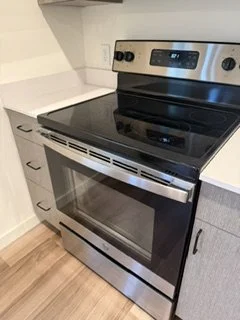 Stainless steel oven and stove with a digital display and control knobs, situated in a kitchen with light-colored countertops and cabinets.