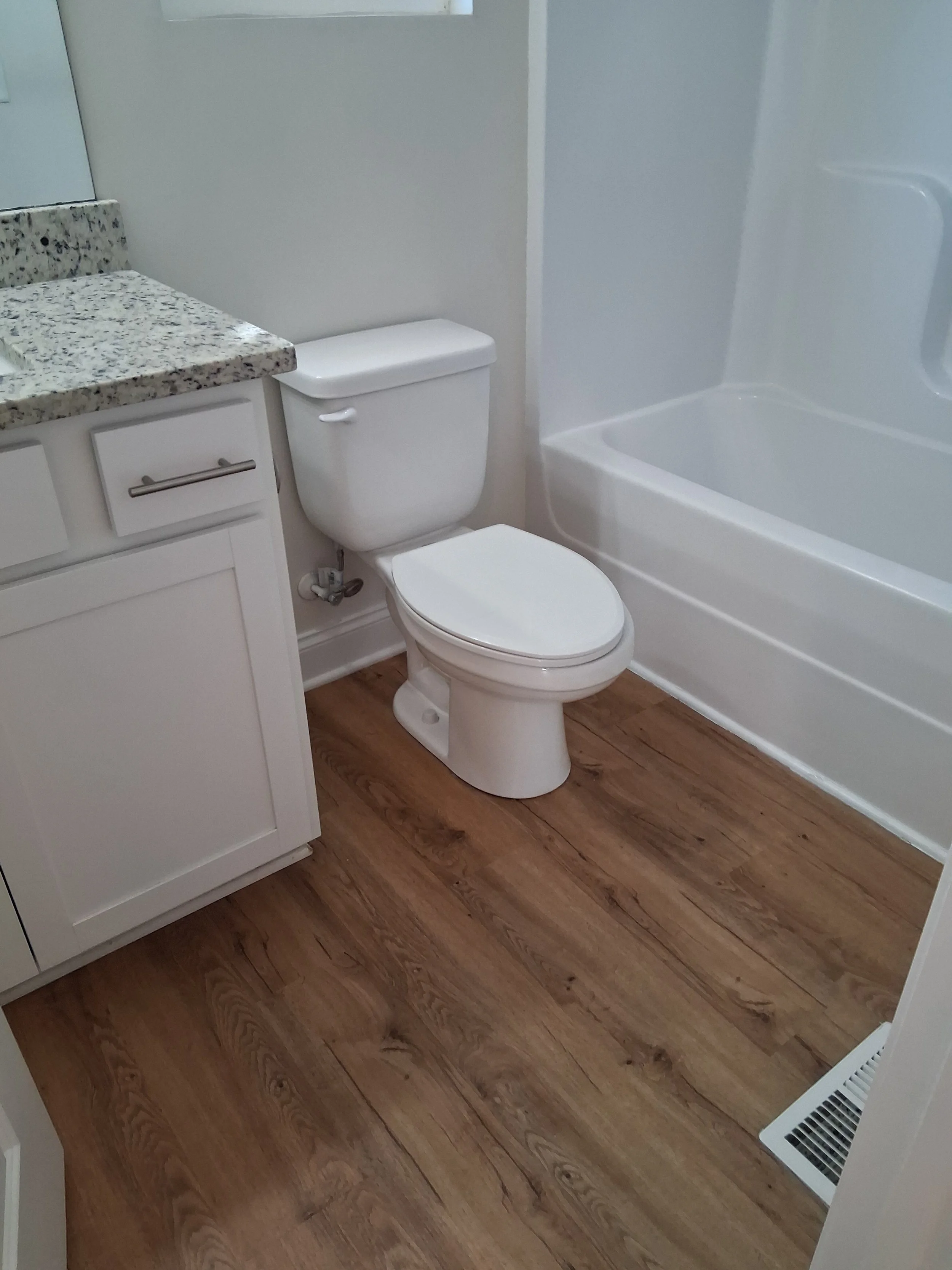 A small bathroom with a white toilet, a white vanity with a granite countertop, and a bathtub with a white surround. The floor has wooden planks, and there is a baseboard heater at the bottom right corner.