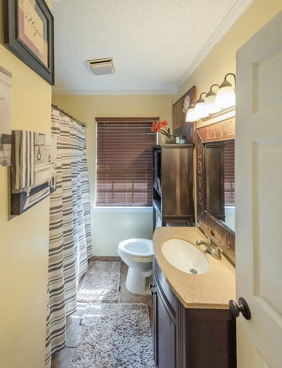 A small bathroom with yellow walls, a shower with striped curtain, a toilet, a vanity with a sink, a mirror with 'LOVE' carved on top, a light fixture with four bulbs, a window with brown blinds, a wooden cabinet, and decorating items including a vas