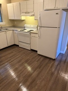 Empty kitchen with white cabinets, refrigerator, stove, and microwave, wooden floor, and yellow backsplash lighting.