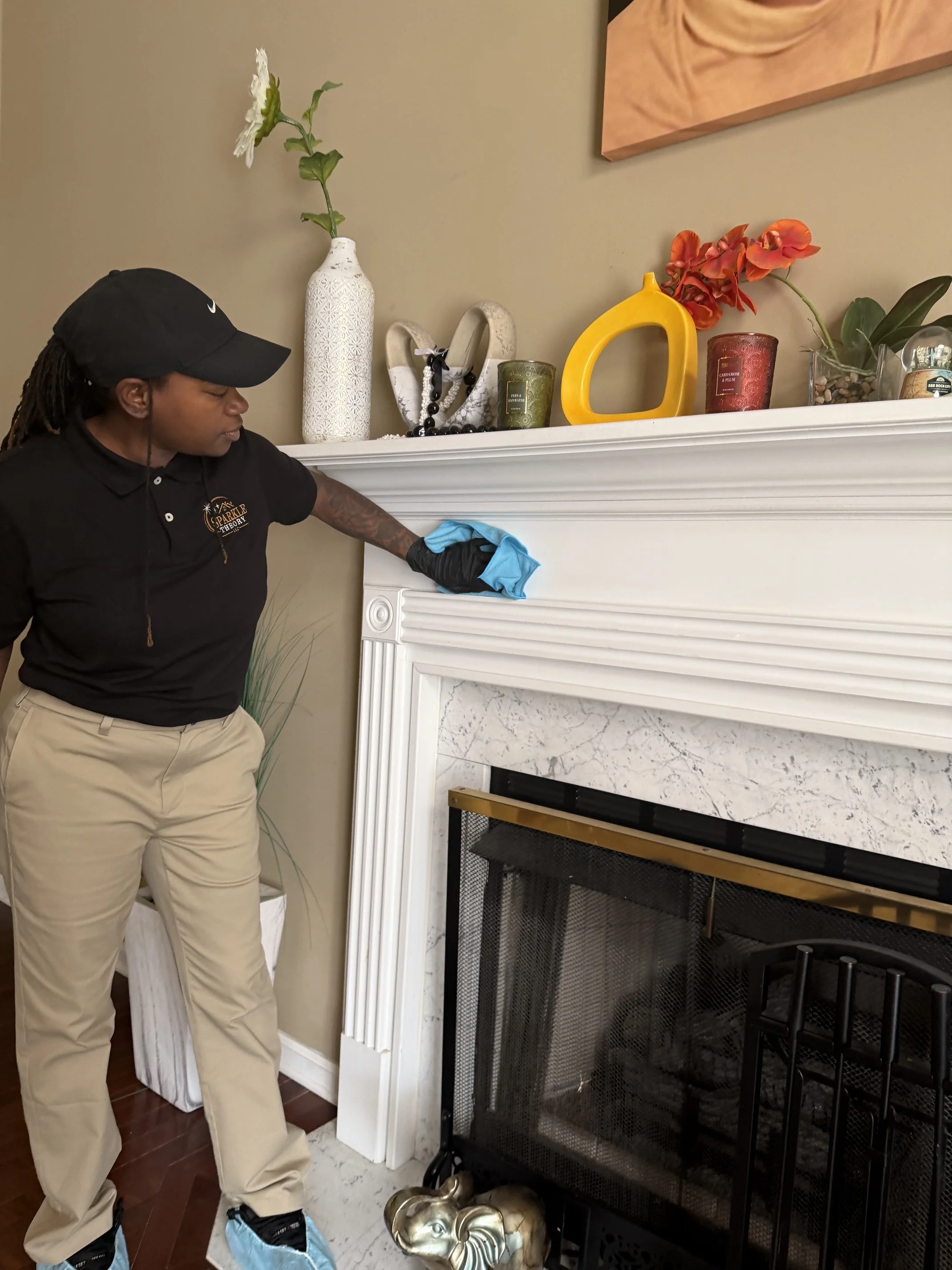 Person cleaning above fireplace with a cloth, wearing a black cap and beige pants, in a living room with decorative items on the mantel including a white vase with a flower, colorful glass candle holders, and a yellow decorative piece.