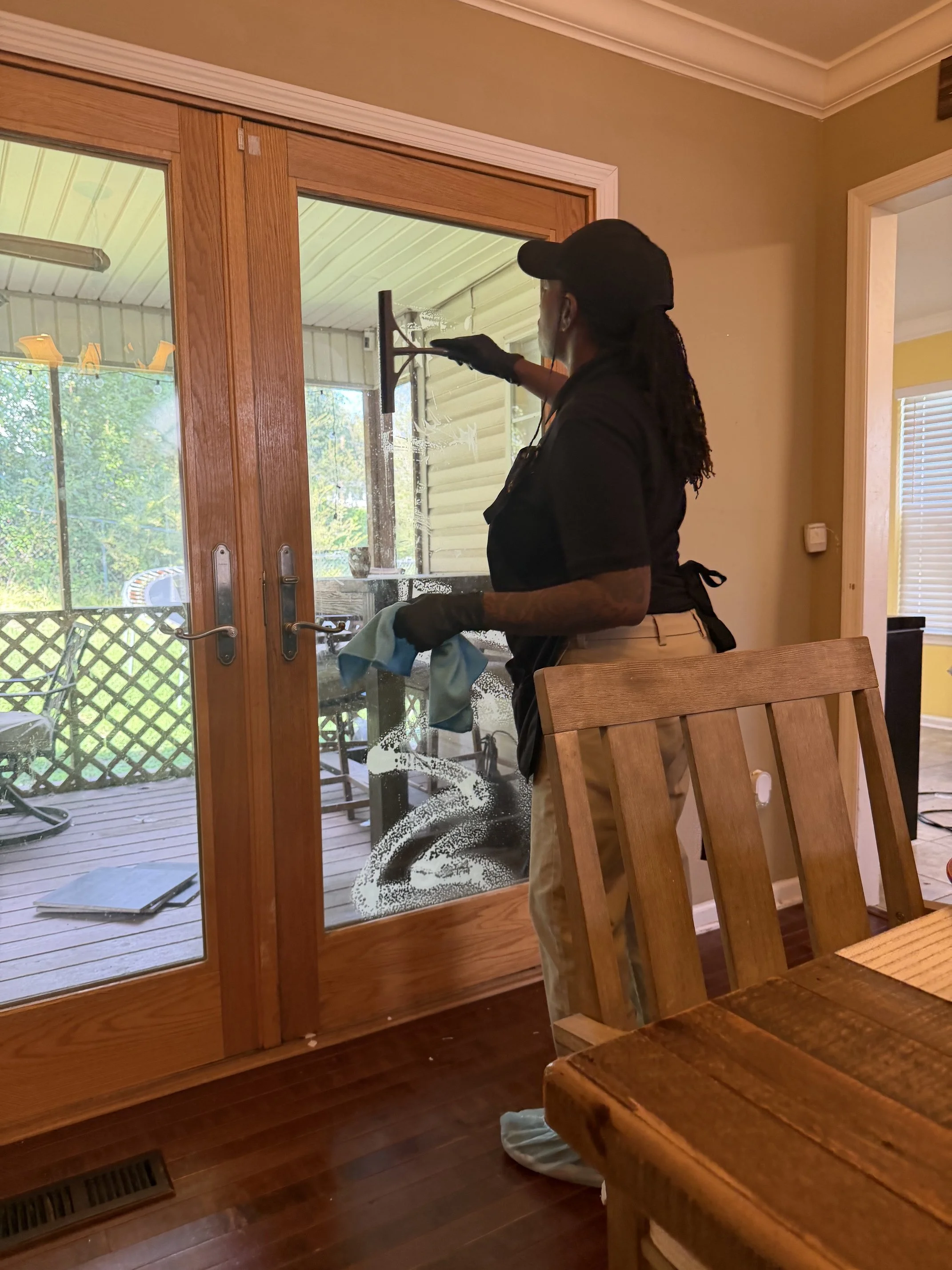 Indoor scene of a woman cleaning the glass sliding door from the inside, using a squeegee. She is wearing a black cap, black gloves, black shirt, and tan pants. The room has a wooden table and chairs, with a view of the outdoor patio with chairs and 