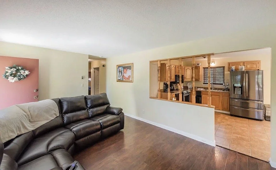 Living room with a black leather sofa, a wall with a pink accent and floral decor, a wall with framed artwork, wood flooring, opening into a kitchen with wooden cabinets, stainless steel appliances, and tiled floor.