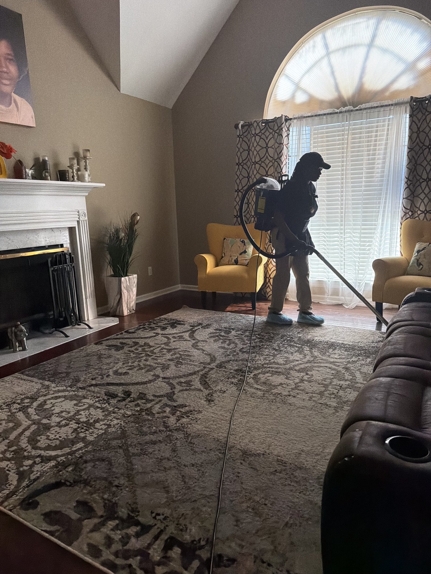 Person cleaning the living room carpet with a vacuum cleaner, standing near a yellow armchair with a pillow, in front of a large window with blinds and patterned curtains, with a fireplace to the left and a patterned area rug on the floor.