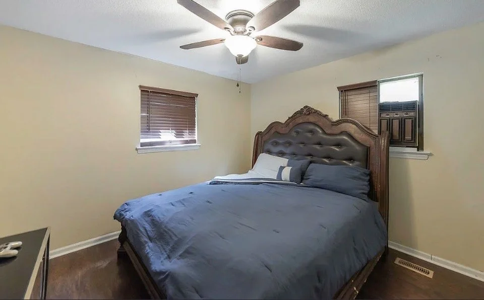 A bedroom with a wooden bed frame with a tufted headboard, dark blue bedding, and two small windows with blinds, beige walls, a ceiling fan with lights, and a small vent on the floor.