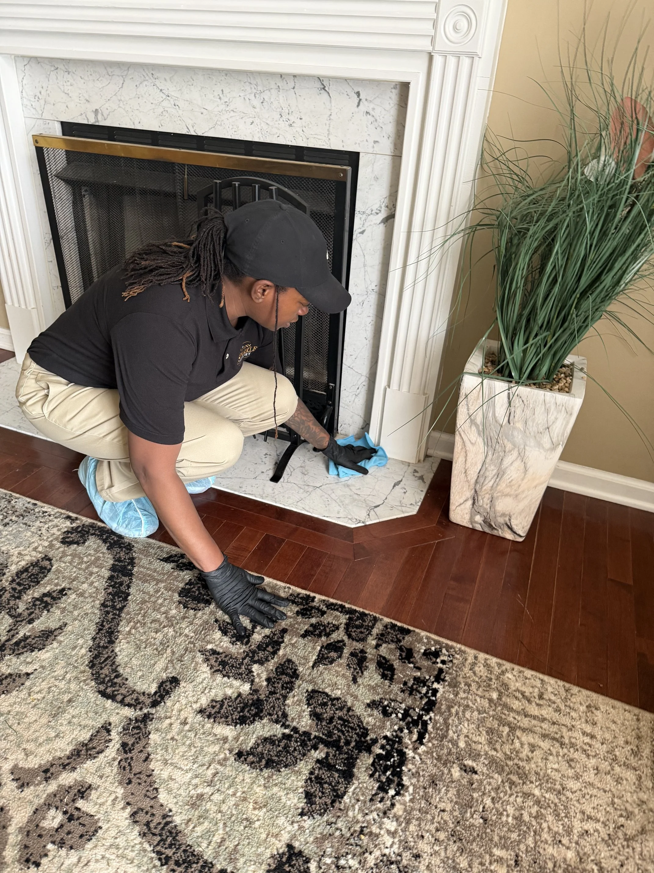 A person wearing a black cap, sunglasses, and black gloves is cleaning or inspecting a fireplace area in a home, crouching near a patterned rug and a marble fireplace with a large potted plant nearby.