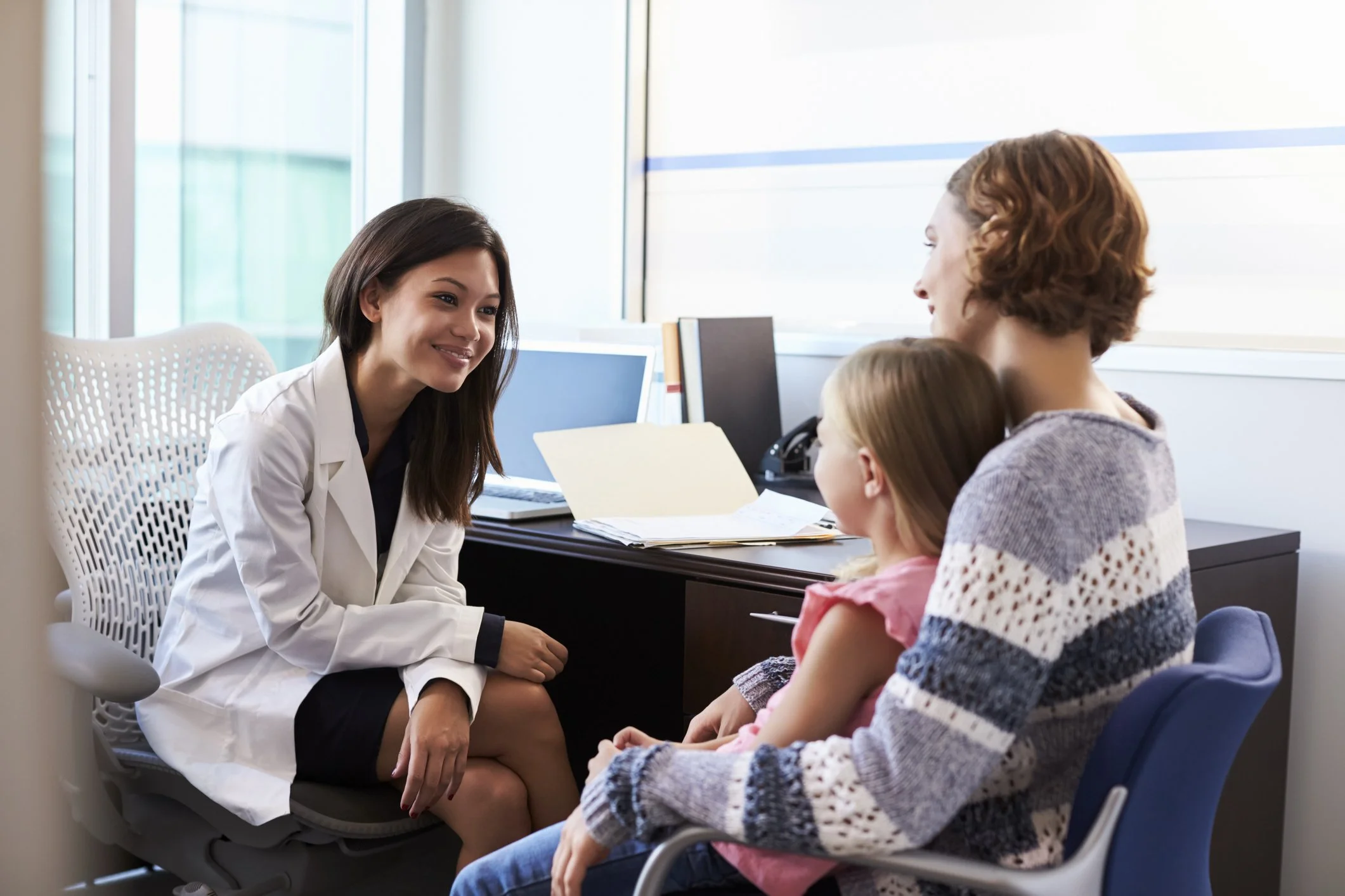 A medical professional, woman with long dark hair wearing a white lab coat, is sitting and engaging in a conversation with two young girls, one with blonde hair wearing a pink top and the other with brown hair wearing a sweater. They are in a bright, modern medical office with large windows.