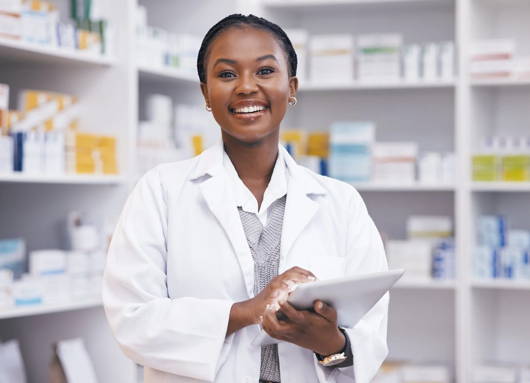 A smiling woman in a white lab coat holding a tablet in a pharmacy with shelves of medications behind her.