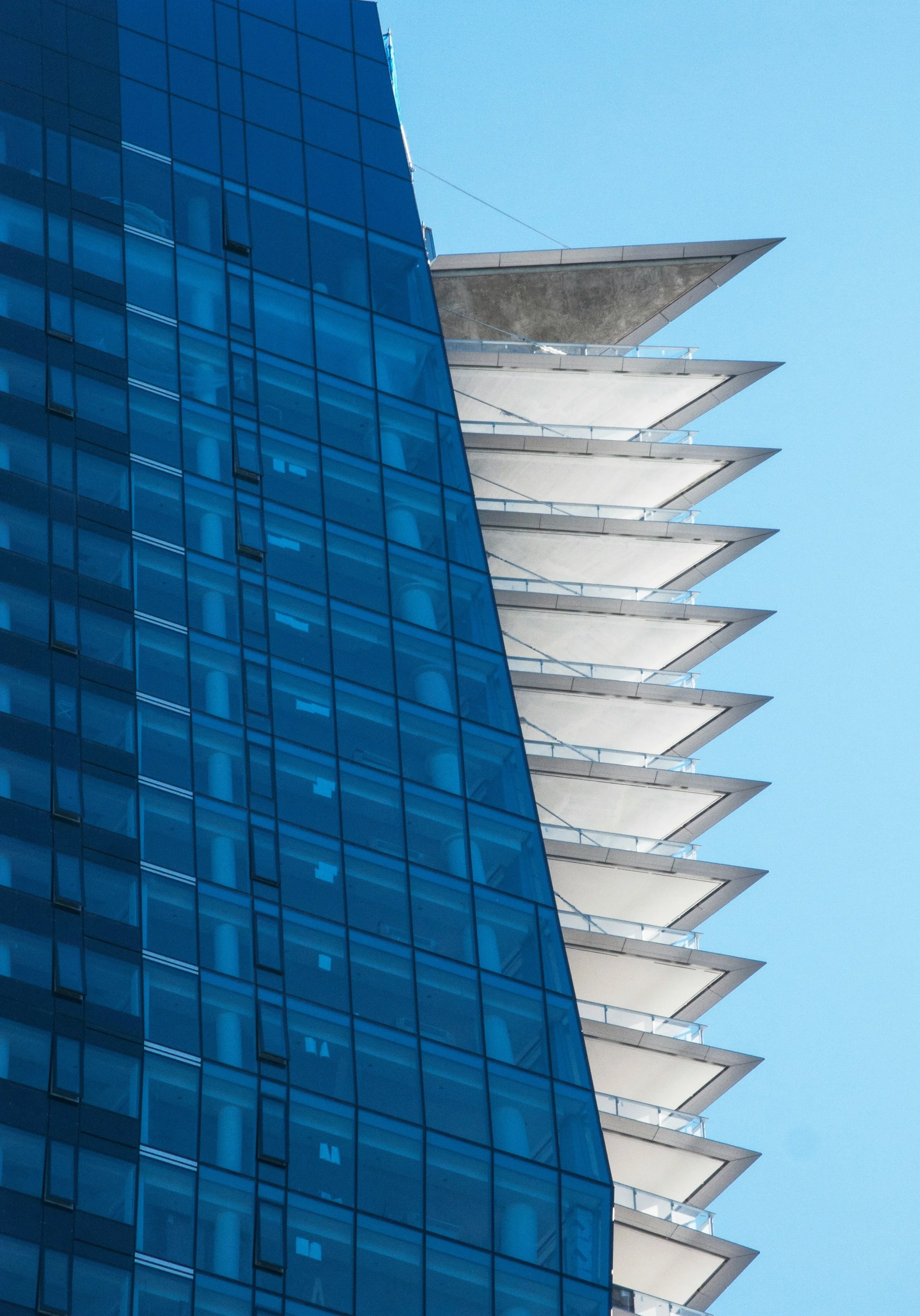 Close-up of a modern building with blue reflective glass windows on the left and concrete balconies with pointed edges on the right, set against a clear blue sky.