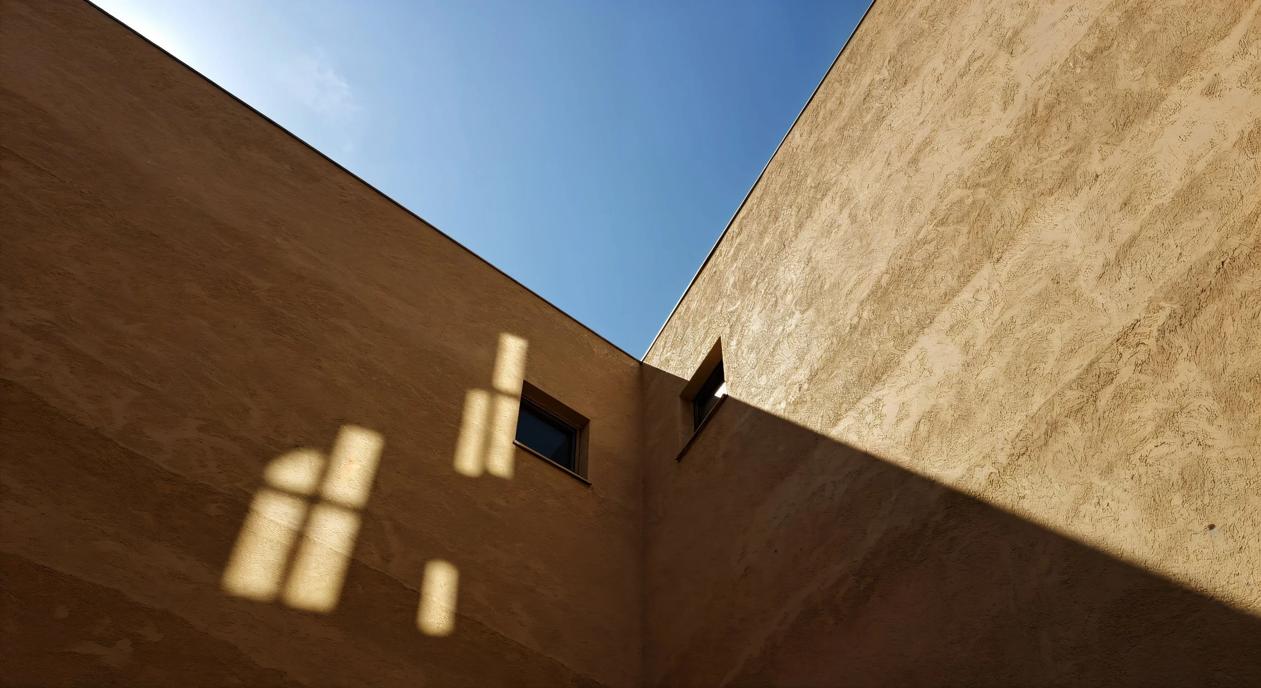 Looking up at two beige stucco building walls meeting at a corner with a small window in one wall and sunlight casting shadows.