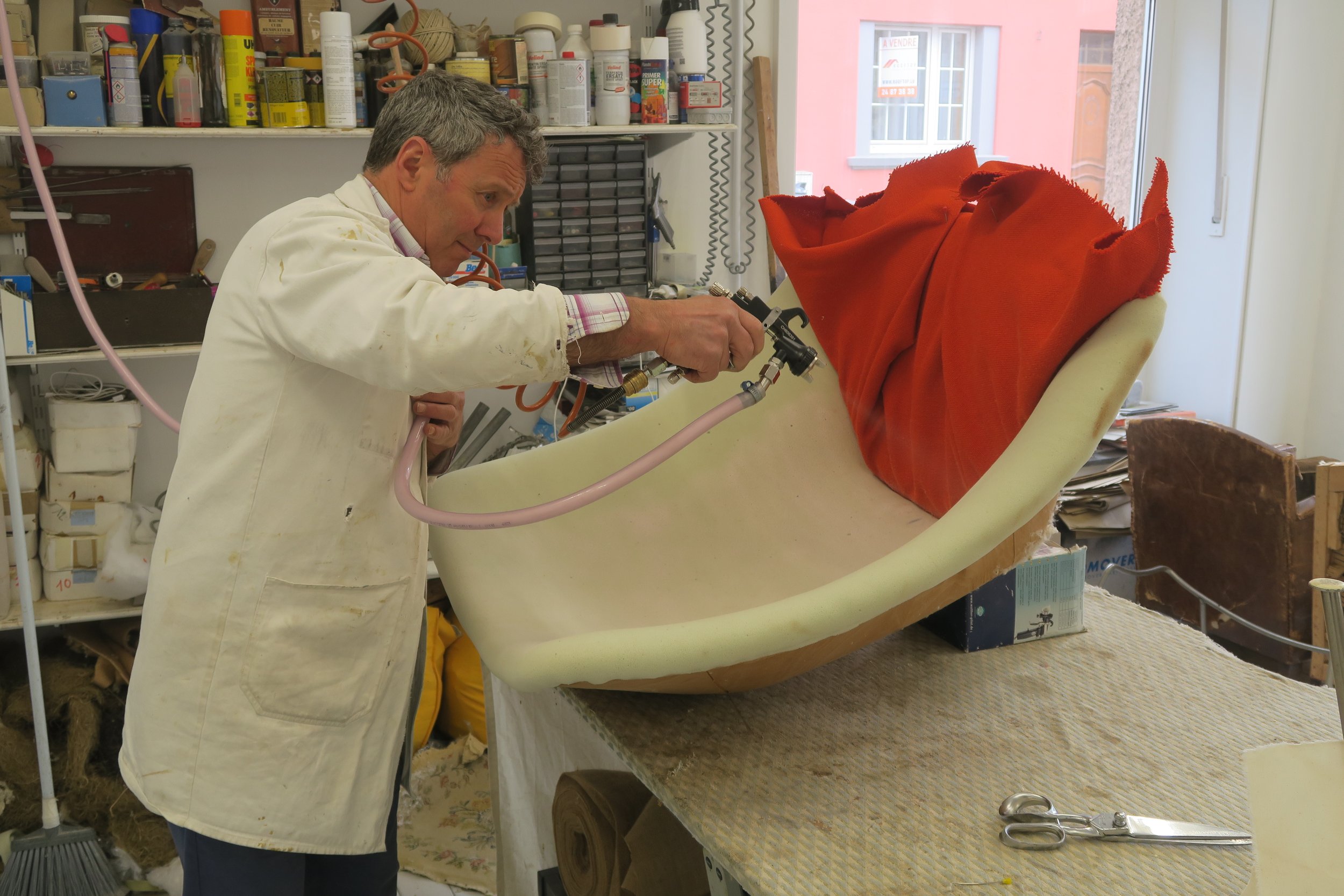 Un homme en blouse de laboratoire utilisant un pistolet pour spray un objet de grande taille recouvert d'une pièce en tissu orange, dans un atelier ou un laboratoire.